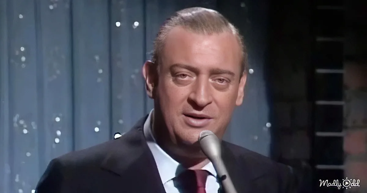A man in a suit and tie speaks into a microphone on stage, with a backdrop of blue curtains and lights.