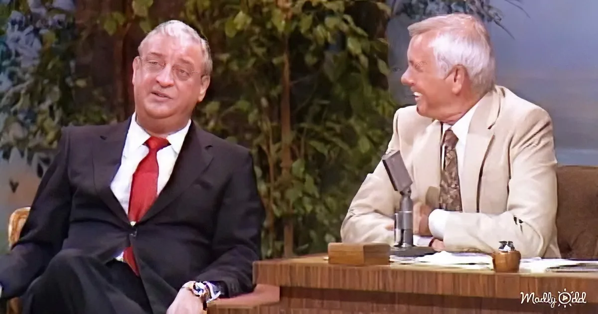 Two men in suits, one in black with red tie, seated at a desk with a microphone, engaged in conversation.
