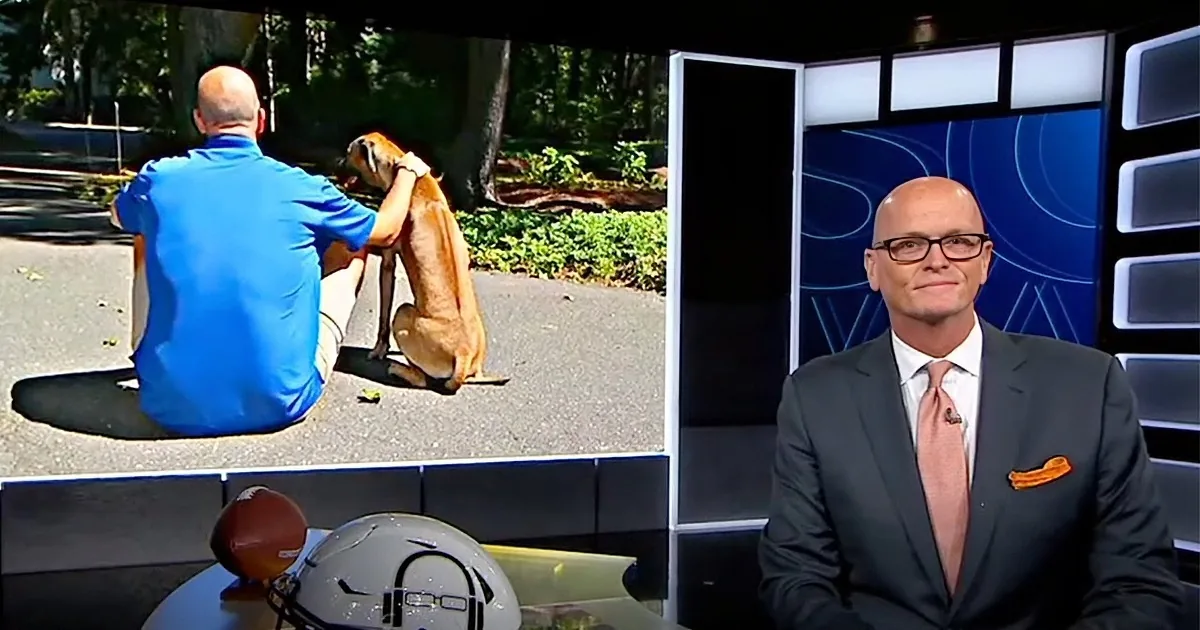 A man in a suit and glasses sits on a set, while another man in a blue shirt pets a dog outdoors.