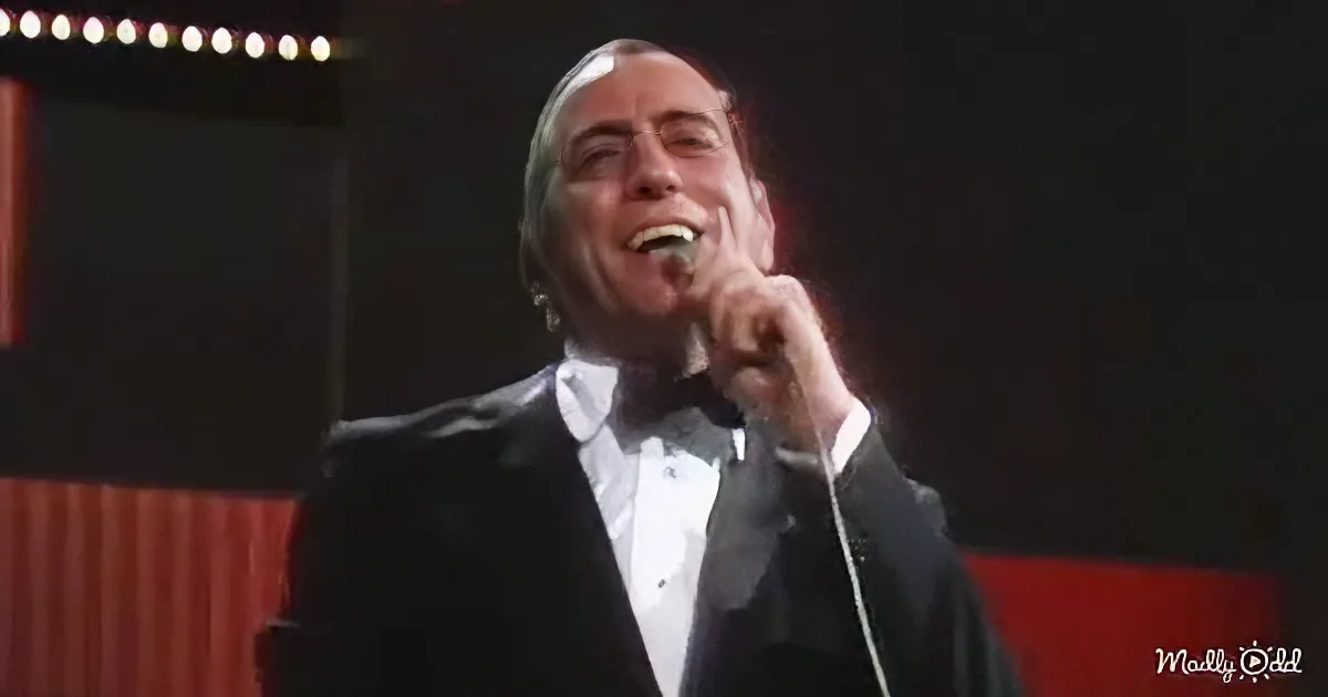 A man in a tuxedo sings into a microphone on stage, illuminated by stage lights.