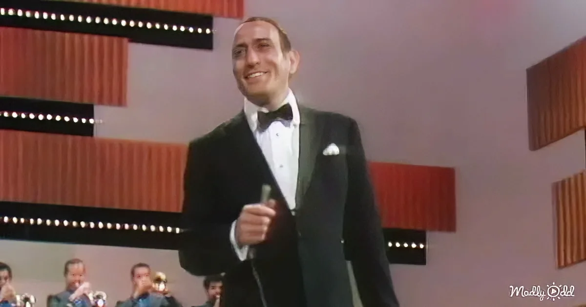 A man in a tuxedo smiles while holding a microphone, standing in front of a band.
