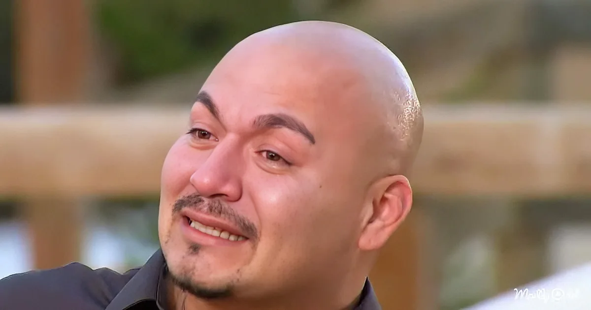 A bald man with a mustache and goatee, wearing a dark shirt, smiles slightly while looking to the side.