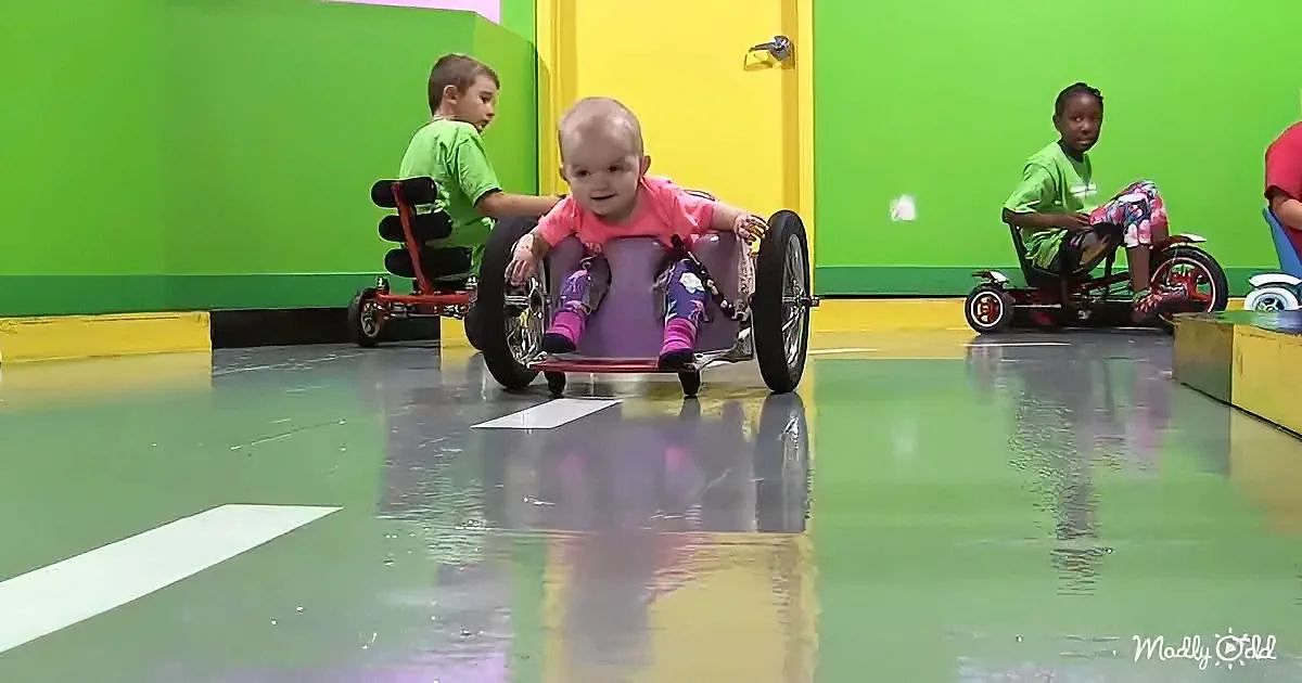 A baby in a purple wheelchair is in the foreground, surrounded by children on tricycles in a colorful indoor play area.