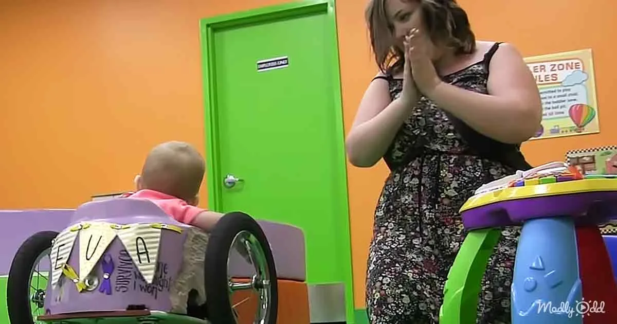 A woman in a floral dress stands near a baby in a walker with a banner, in a colorful room.