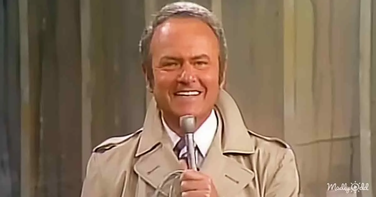 A man in a beige trench coat smiles while holding a microphone, standing against a curtain backdrop.