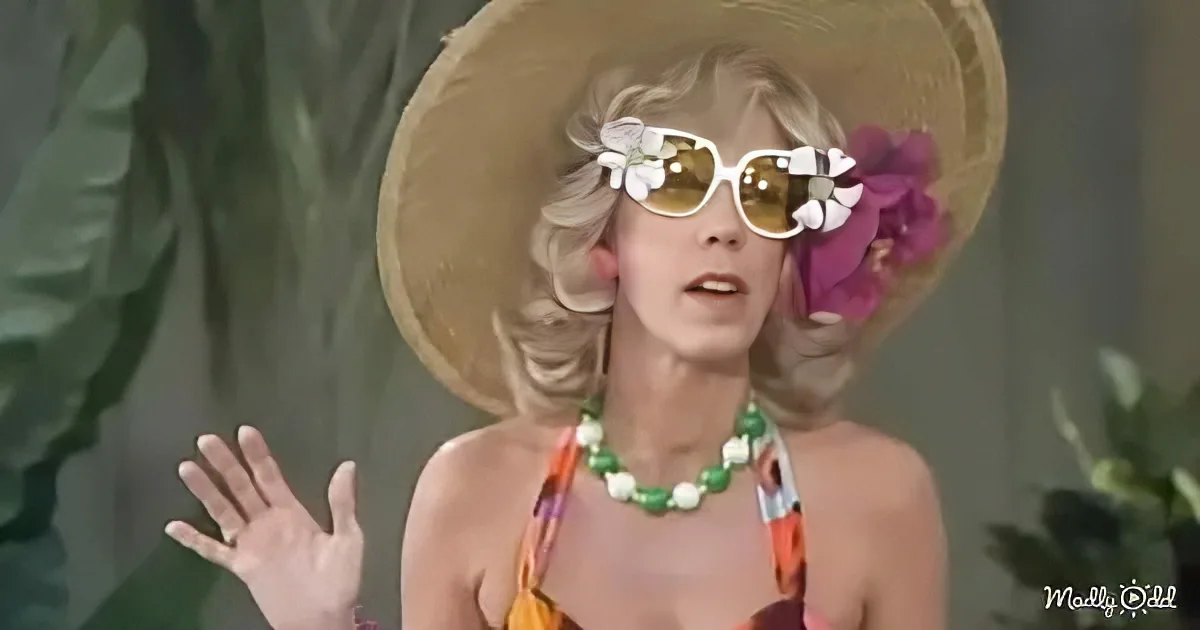 A woman in a colorful outfit, large straw hat, and floral sunglasses gestures with her hand.