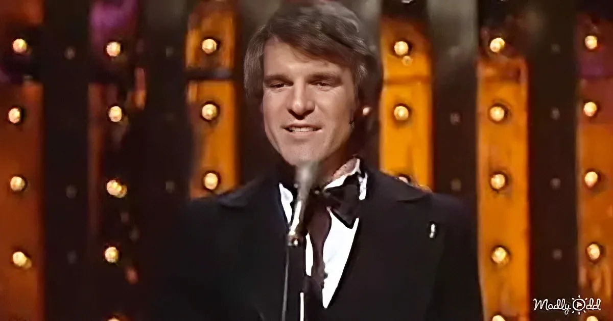 A man in a tuxedo smiles at a microphone on stage with a backdrop of lights.