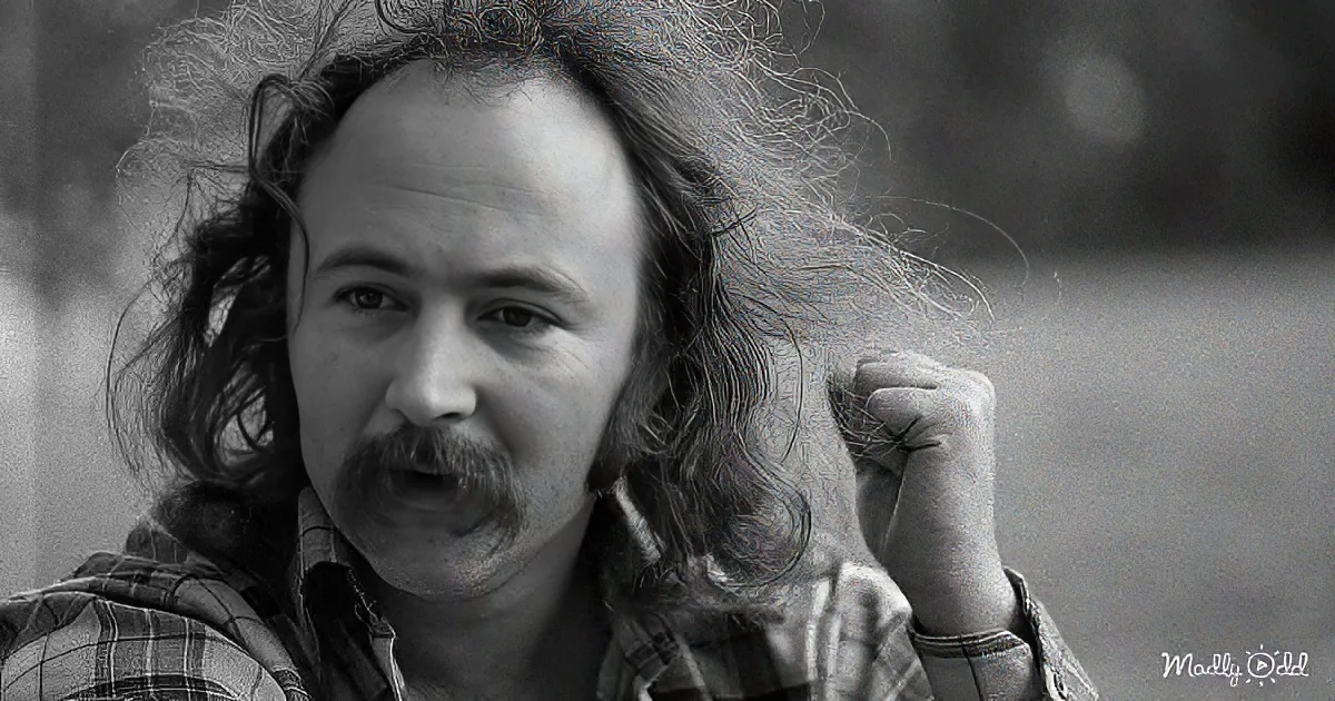 A man with long hair and a mustache, wearing a plaid shirt, holds his hand near his ear.