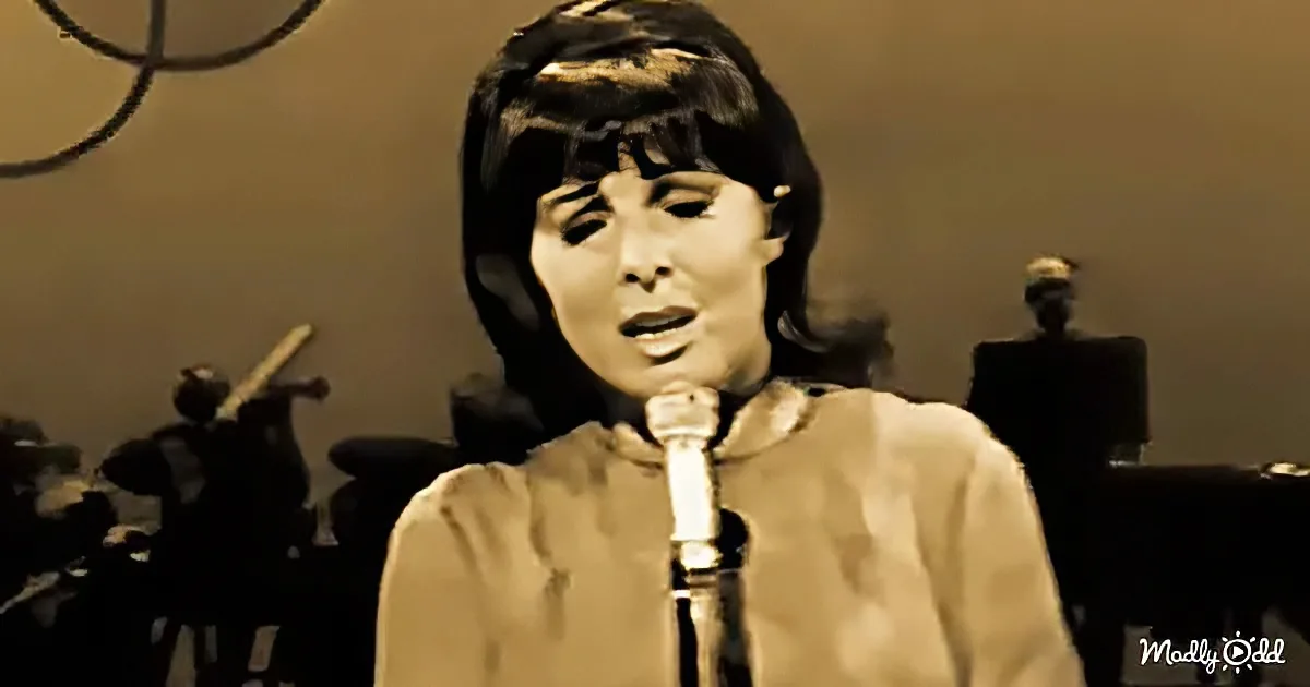 A woman sings into a microphone, wearing a light-colored top, with a band in the background.