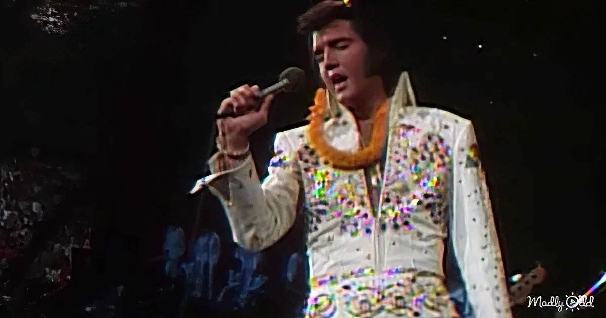 Elvis Presley in a white sequined jumpsuit singing into a microphone.