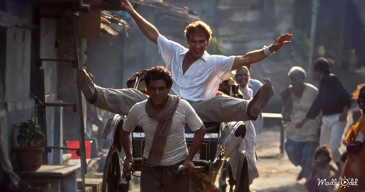 A man in a white shirt is joyfully riding a rickshaw, arms outstretched, in a bustling street scene.