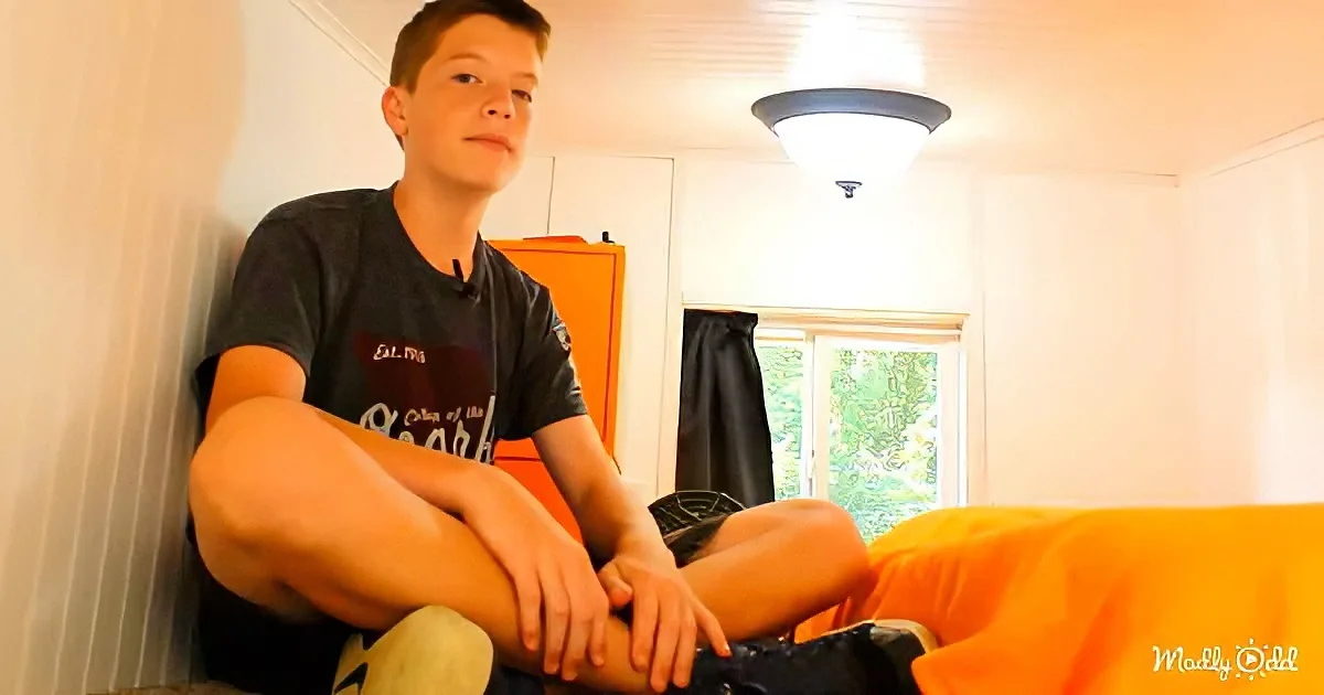A young boy sits on a bed, wearing a dark t-shirt, with his legs crossed, in a room with a window and orange furniture.