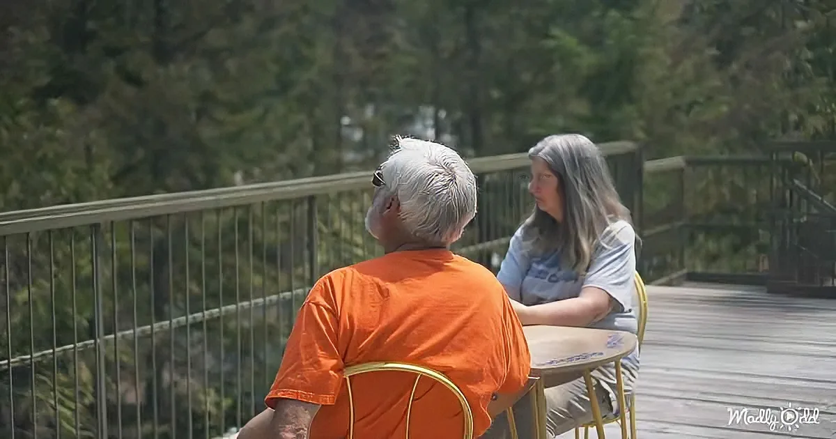 An elderly man in an orange shirt sits at a table with a woman, both on a wooden bridge surrounded by trees.