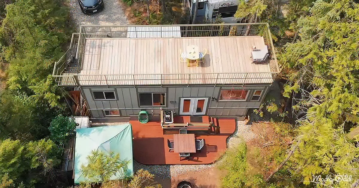 A small house with a wooden deck, outdoor furniture, and a green tarp-covered area surrounded by trees.