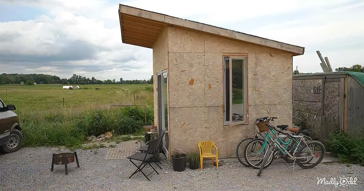 A small, unfinished cabin with bicycles and chairs outside, set in a rural field with a cloudy sky.
