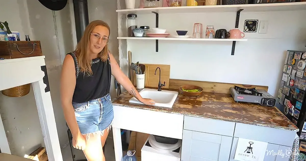 A woman stands in a rustic kitchen with a white sink, wooden countertop, and shelves holding various kitchen items.