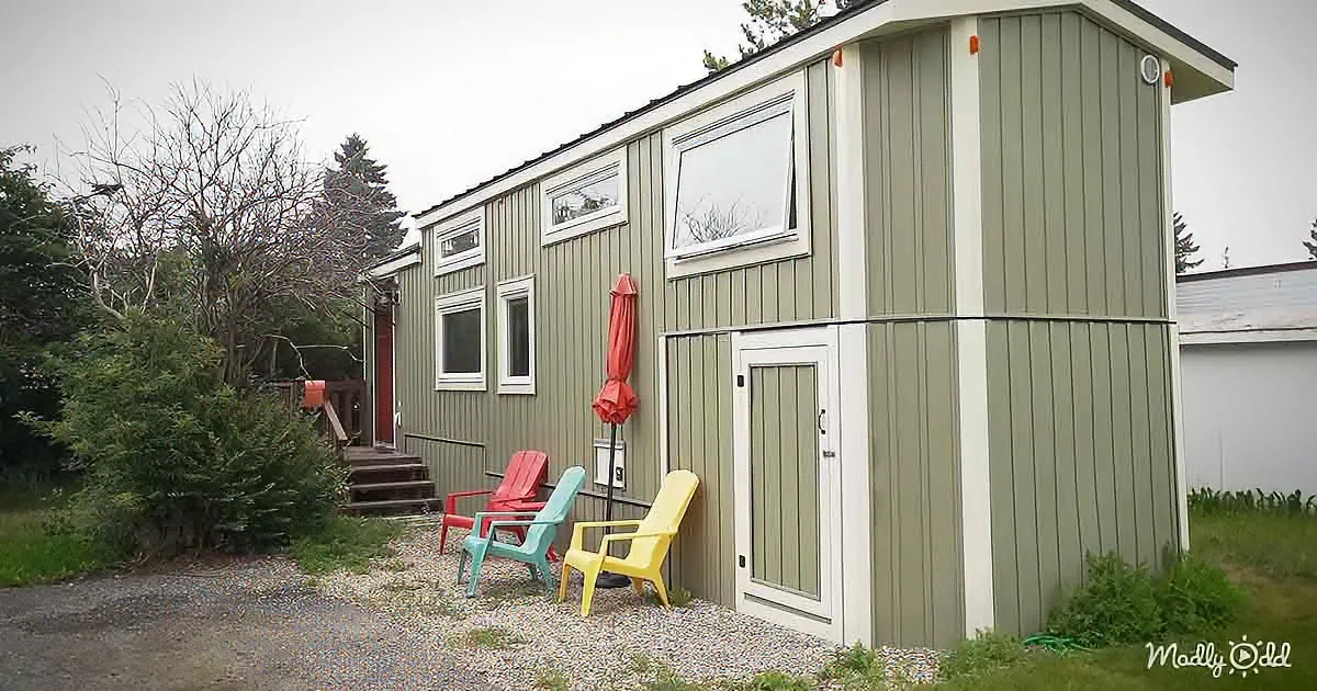 A small, green, wooden cabin with colorful chairs and an umbrella outside.