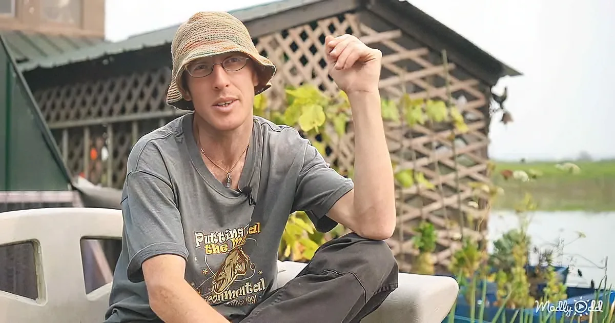 A man wearing a hat and glasses sits outdoors, gesturing with his right hand, with a wooden structure and plants in the...