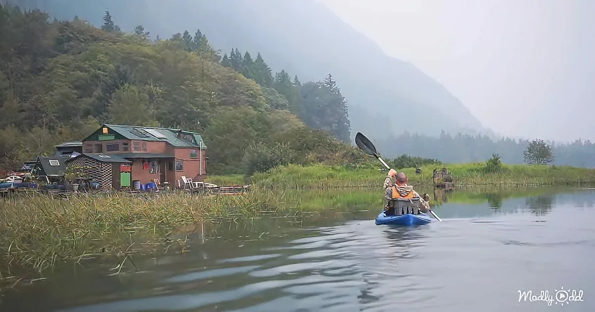 A person paddles a blue kayak on a calm lake near a house with a green roof.