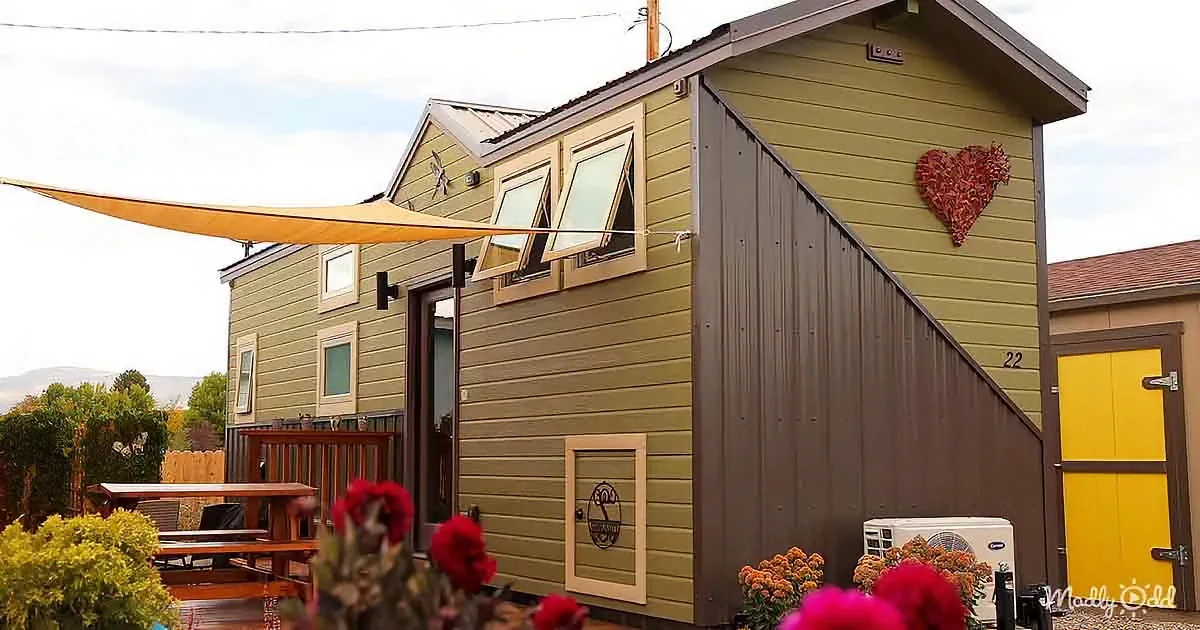 A charming green tiny house with a heart decoration, yellow door, and outdoor seating area.