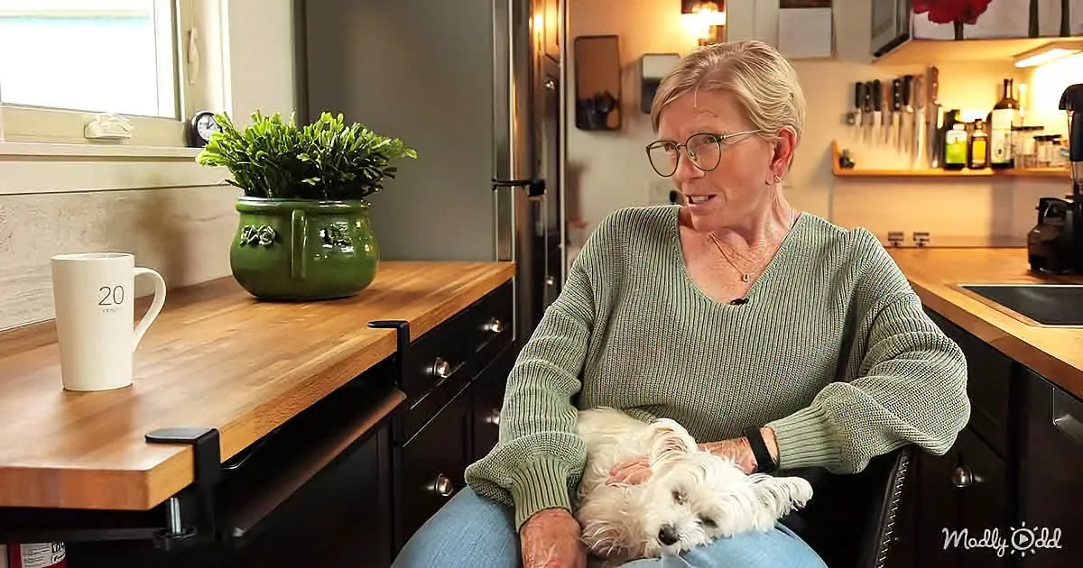 A woman in a green sweater sits in a wheelchair, holding a white dog, with a kitchen counter and green pot plant in the...
