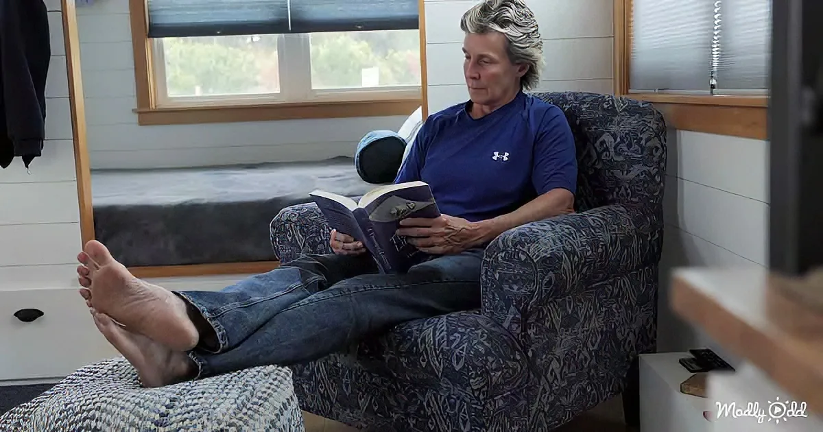 A person sits relaxed in a patterned armchair, reading a book with bare feet up on a footrest.
