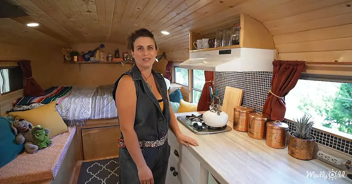 A woman stands in a cozy, compact kitchen with wooden walls, a bed, and a window view of trees.