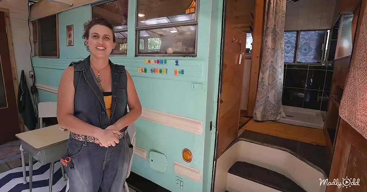 A woman stands in front of a vintage bus with "Welcome to Old School Bus" sign, smiling.