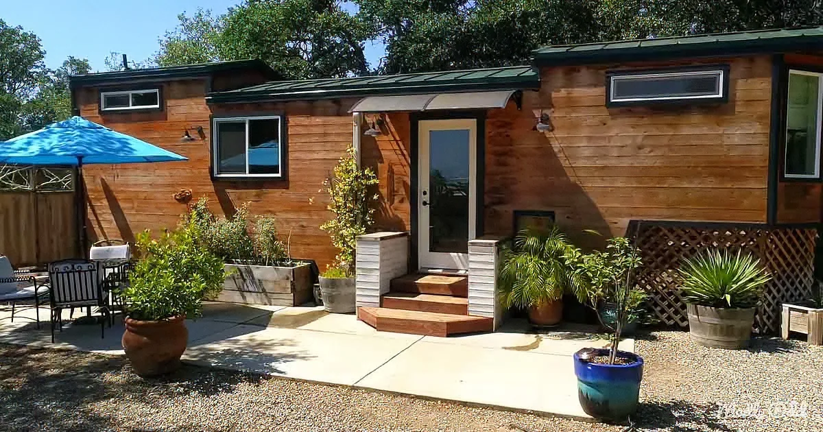 A cozy wooden cabin with a green roof, surrounded by potted plants and a patio area with a blue umbrella.