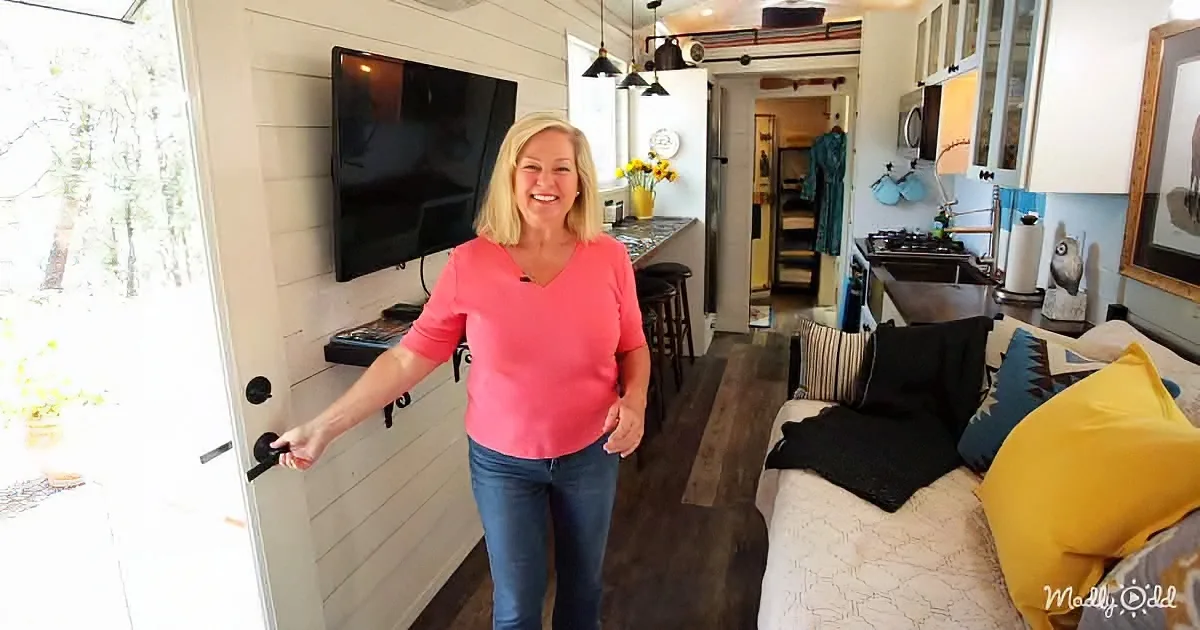 A woman in a pink shirt smiles while holding a door handle in a cozy, well-lit living space with a TV, kitchen, and couch.