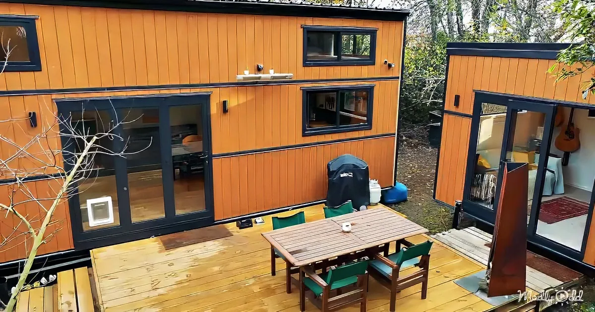 A modern orange tiny house with large windows, a wooden deck, and outdoor seating.
