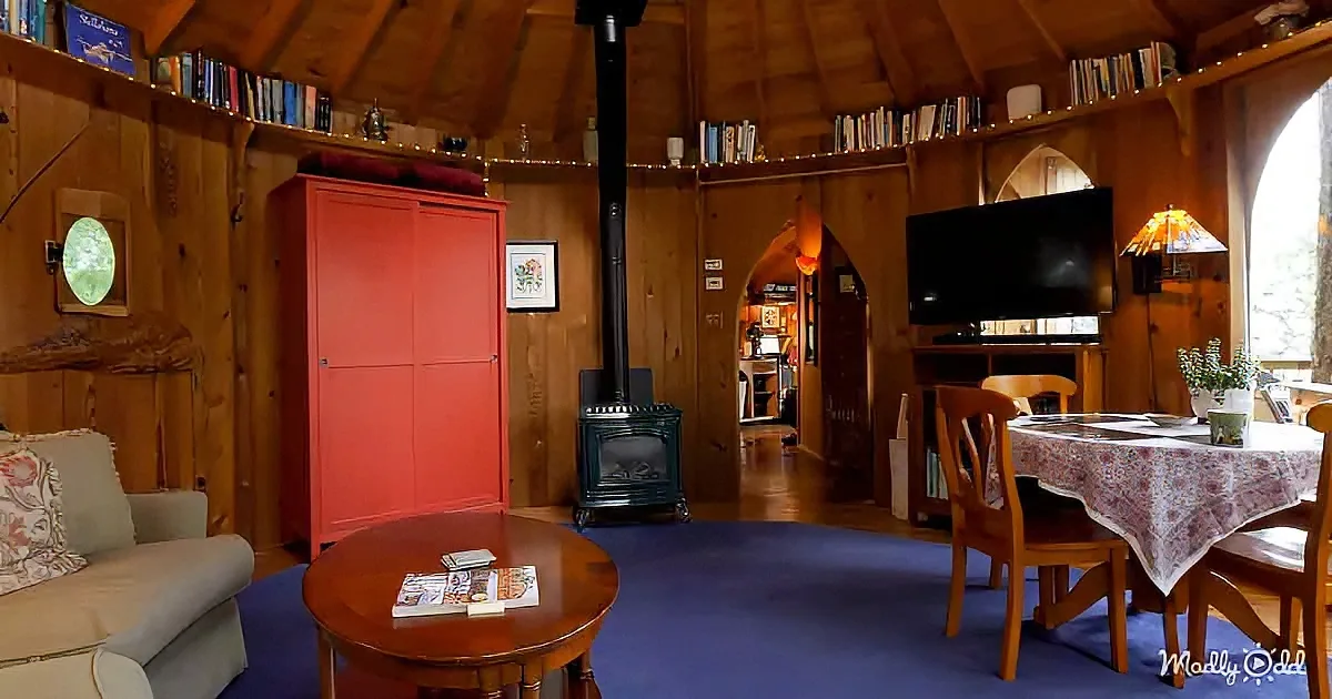A cozy cabin interior with wooden walls, a red cabinet, a wood-burning stove, and a dining area with a floral tablecloth.