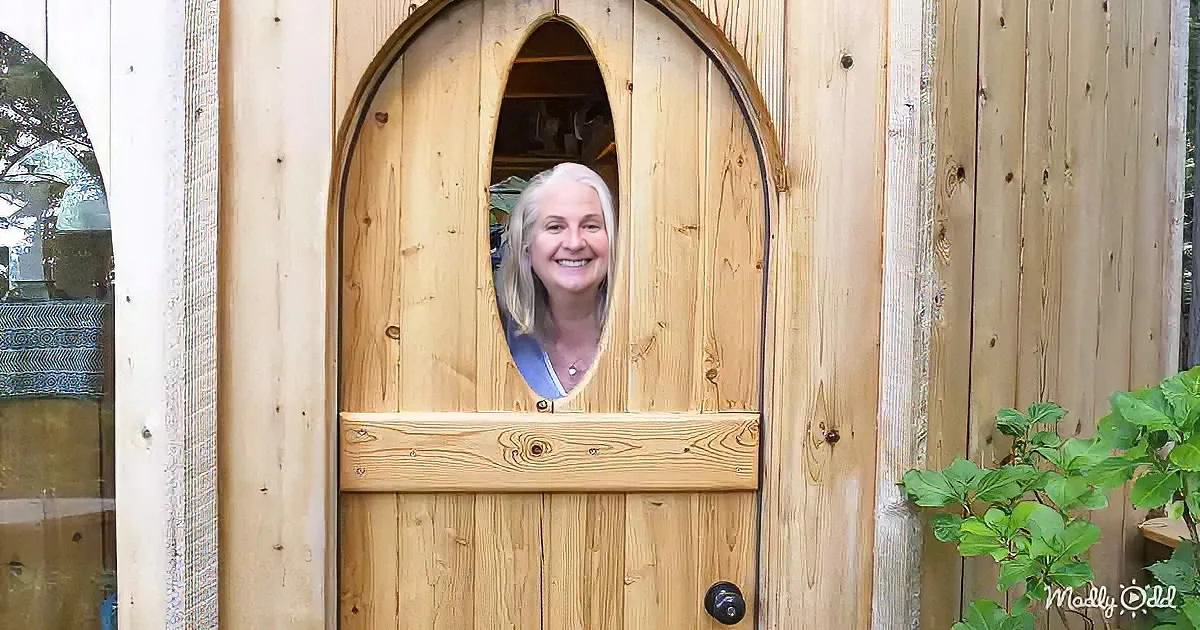 A smiling woman peers through a wooden door with an arched window, surrounded by greenery.