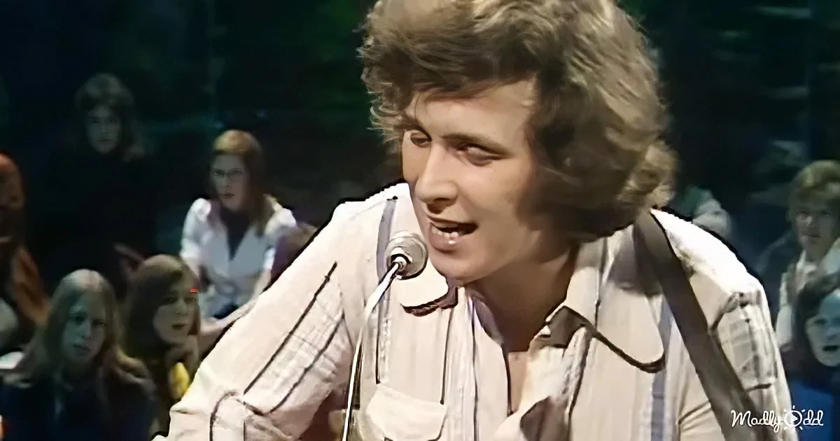 A man with shoulder-length hair sings into a microphone, wearing a white shirt, with a crowd in the background.