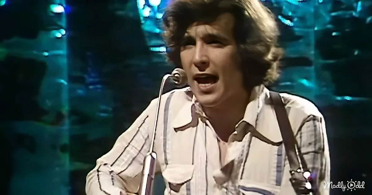 A person in a plaid shirt sings into a microphone on stage with blue lighting.