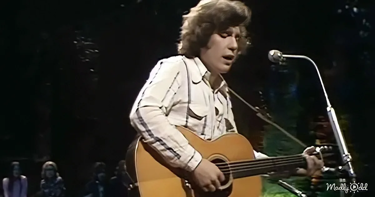 A person with shoulder-length hair plays an acoustic guitar on stage, wearing a white shirt with plaid sleeves.