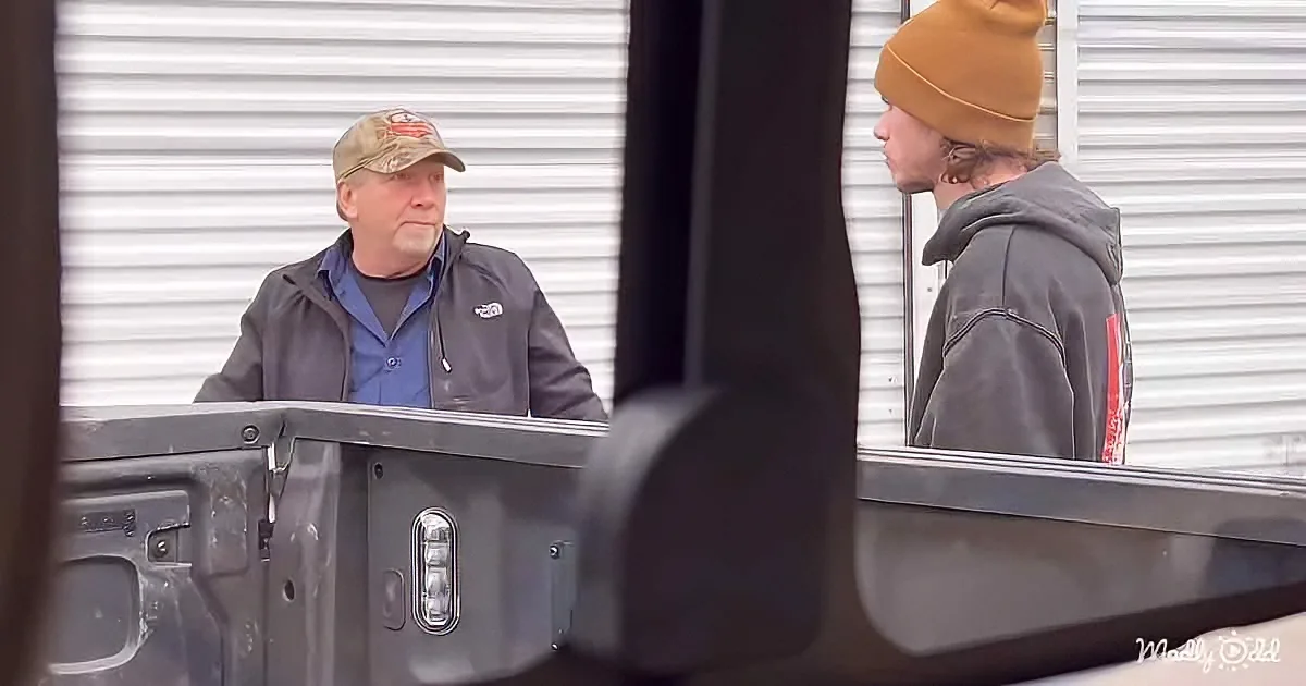 An older man in a cap and jacket stands beside a vehicle, talking to another person in a hoodie and beanie.