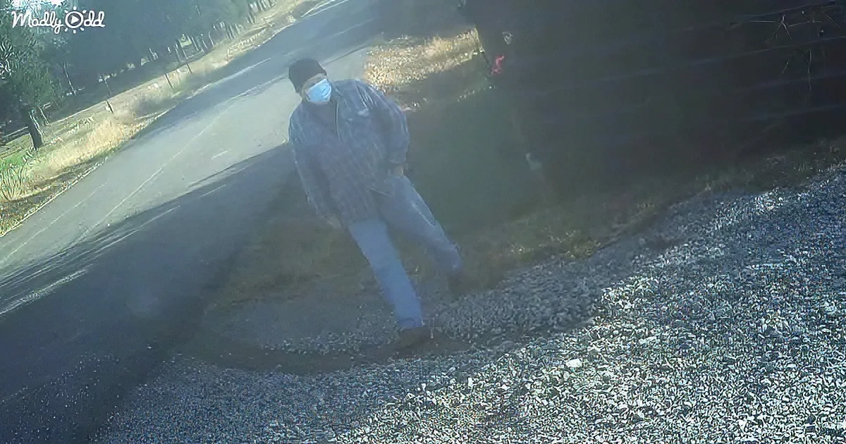A person wearing a mask and dark clothing bends over near a gravel area, possibly picking something up.