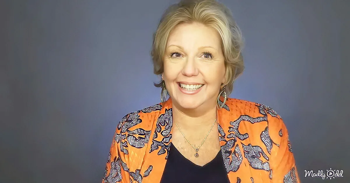 A smiling woman with short blonde hair, wearing an orange patterned jacket and hoop earrings, against a dark background.