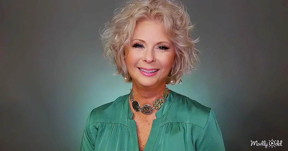 A woman with short, curly blonde hair wearing a green blouse and a statement necklace smiles against a dark background.