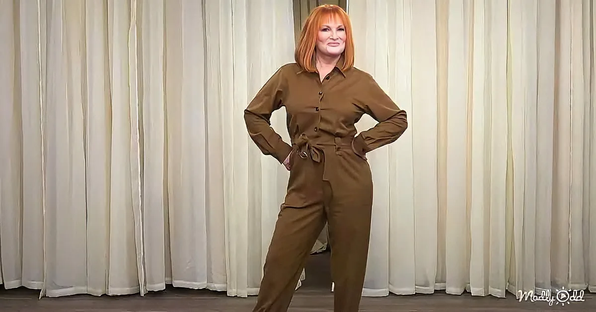 A woman with red hair stands confidently in a brown jumpsuit against a white curtain backdrop.