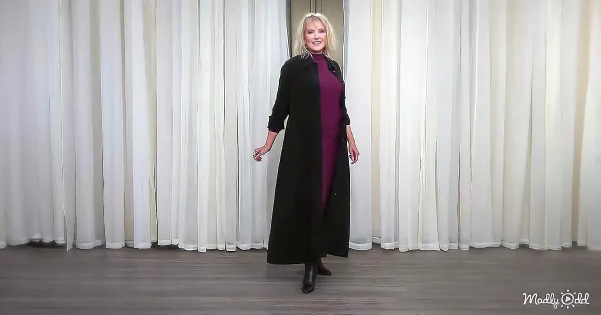 A woman stands in front of a white curtain, wearing a long black coat over a purple dress and black boots.