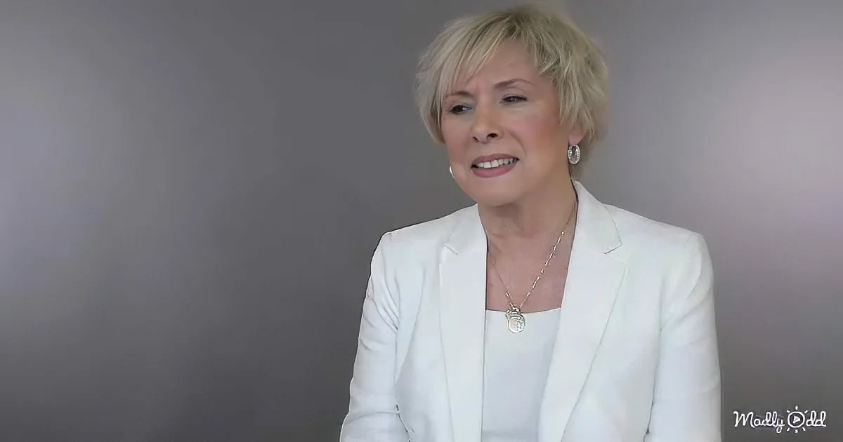 A woman with short blonde hair, wearing a white blazer and necklace, smiles while seated against a gradient gray background.