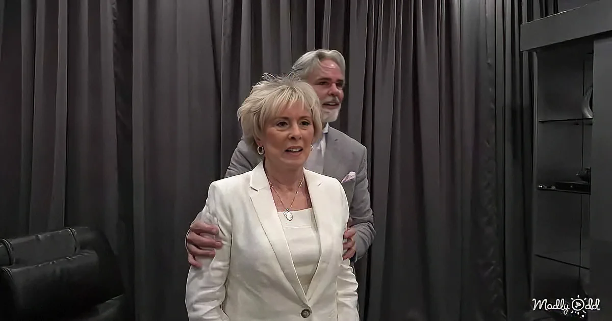 A woman in a white suit jacket stands with a man in a gray suit behind her, both in front of dark curtains.
