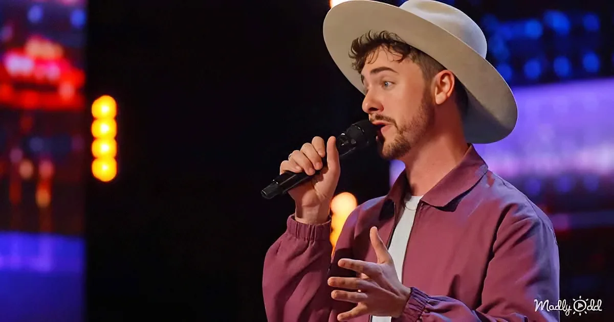 A man in a white hat and maroon shirt sings into a microphone on stage with colorful lights.