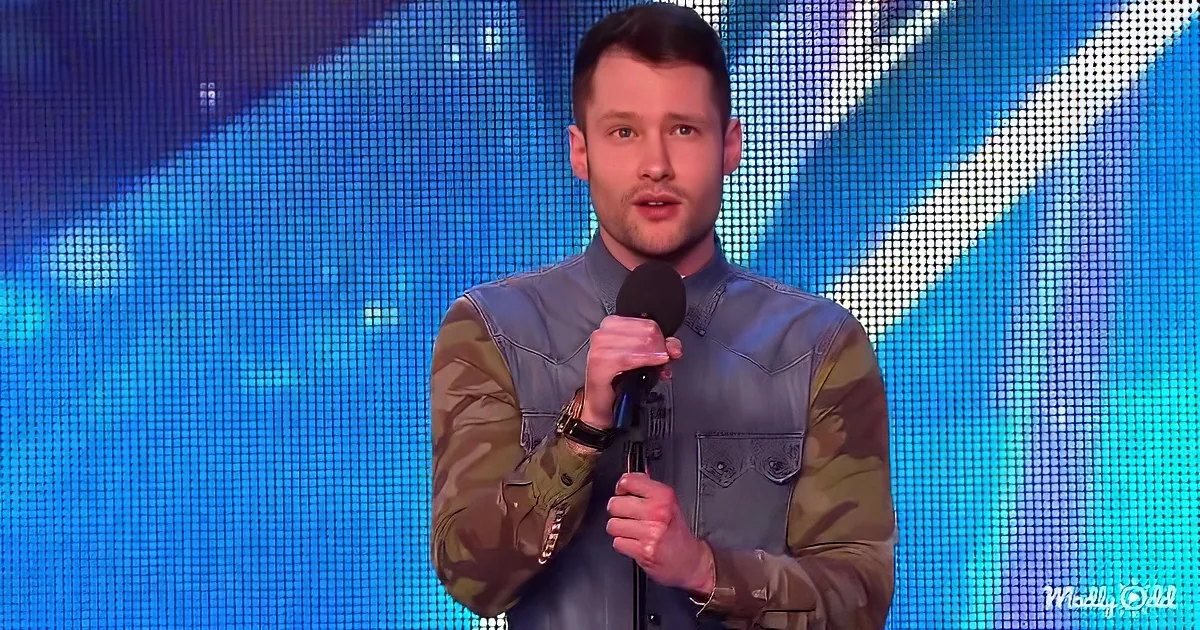 A man in a denim jacket holds a microphone on stage, with a blue LED screen in the background.