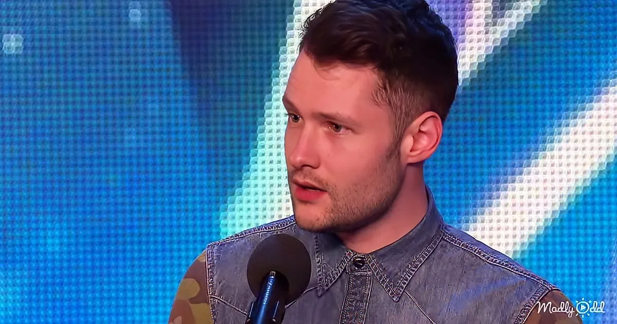A man in a denim shirt speaks into a microphone against a blue, pixelated background.