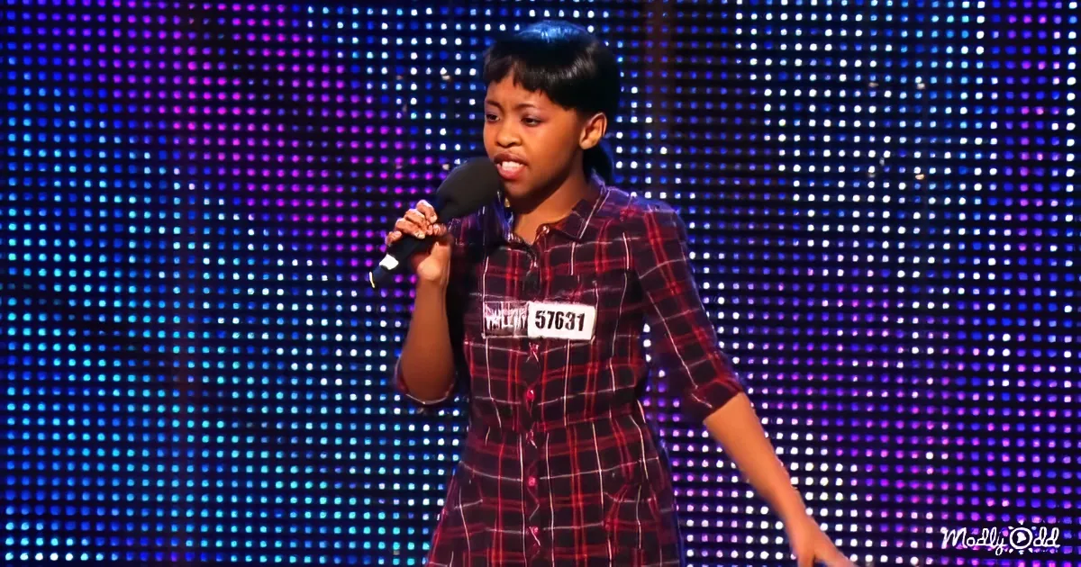 A young girl in a plaid shirt holds a microphone on stage, wearing a name tag.