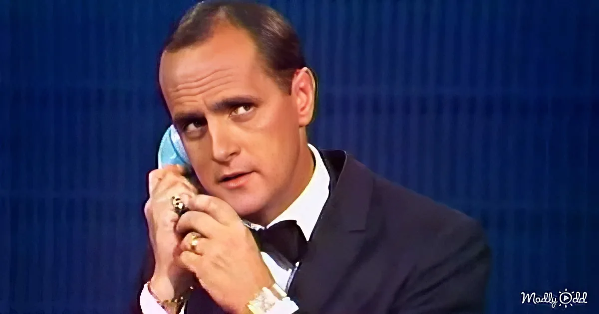 A man in a suit and bow tie holds a phone to his ear, looking serious against a blue background.
