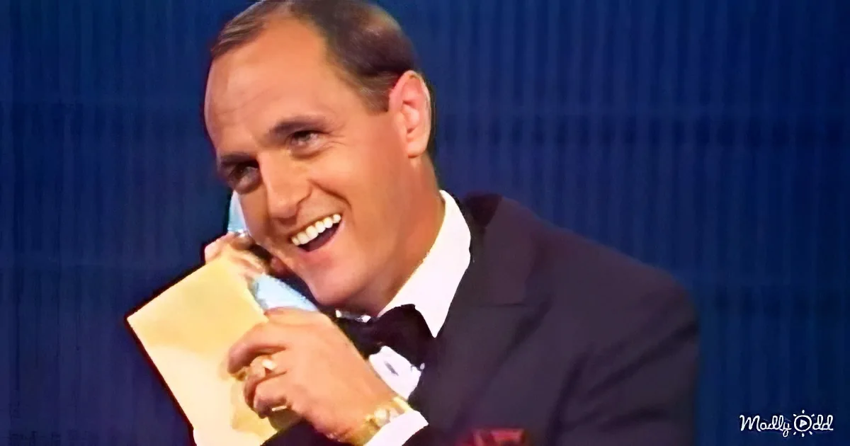 A man in a suit and bow tie smiles while holding a yellow envelope.